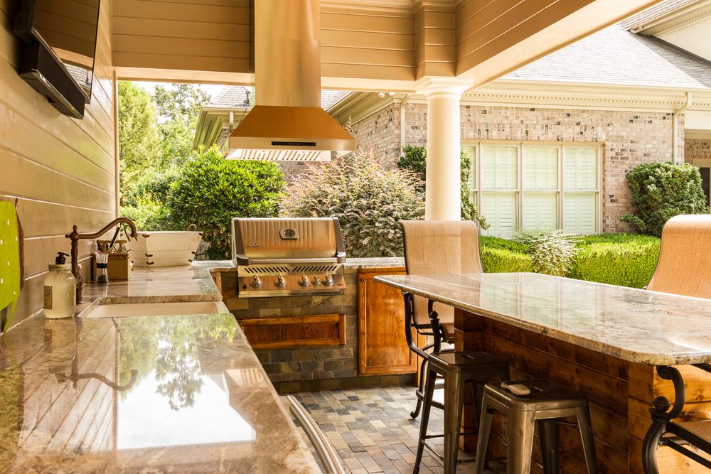 Outdoor kitchen with marble countertops, a grill under a range hood, bar stools and a sink